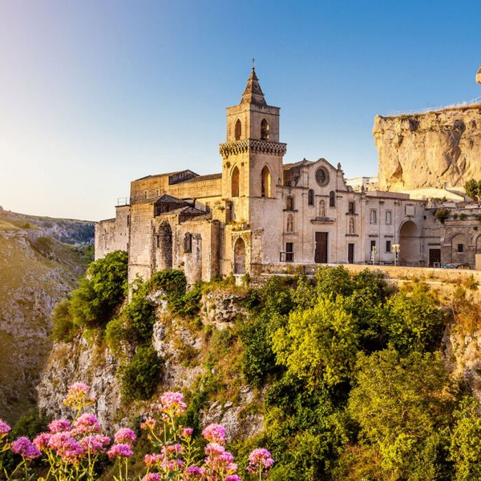 Vista panoramica di un'antica chiesa su una scogliera con vegetazione e fiori in primo piano.